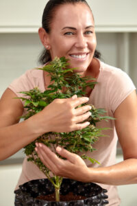 A smiling woman holding and caring for a healthy homegrown cannabis plant in a pot